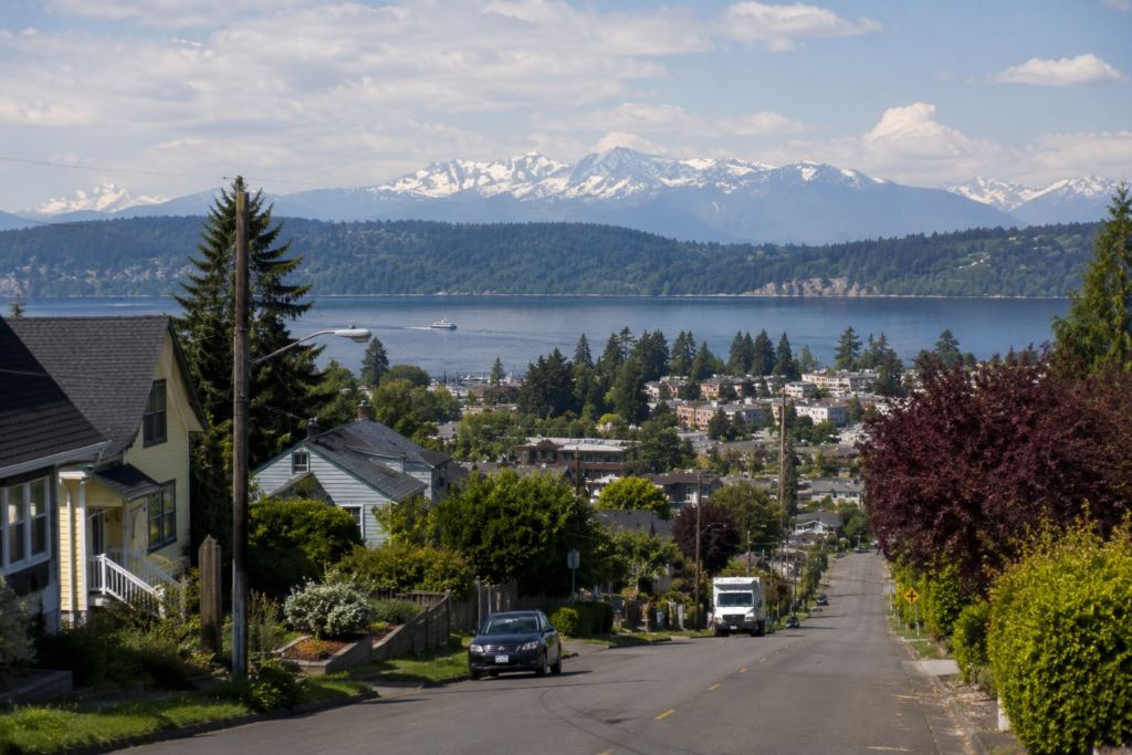 Rua residencial em declive mostrando o limite entre bairros e cidades na região de Puget Sound, com casas, árvores, água e montanhas ao fundo em um dia claro.