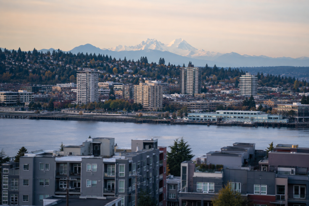 Vista panorâmica da área metropolitana de Seattle com bairros urbanos, edifícios residenciais, trecho de água e montanhas ao fundo, destacando a integração entre diferentes cidades da região de Puget Sound.