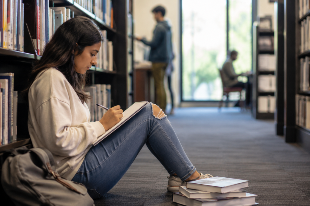 Estudante sentada no chão entre estantes de uma biblioteca escreve em um caderno, com livros ao lado e outros estudantes desfocados ao fundo.