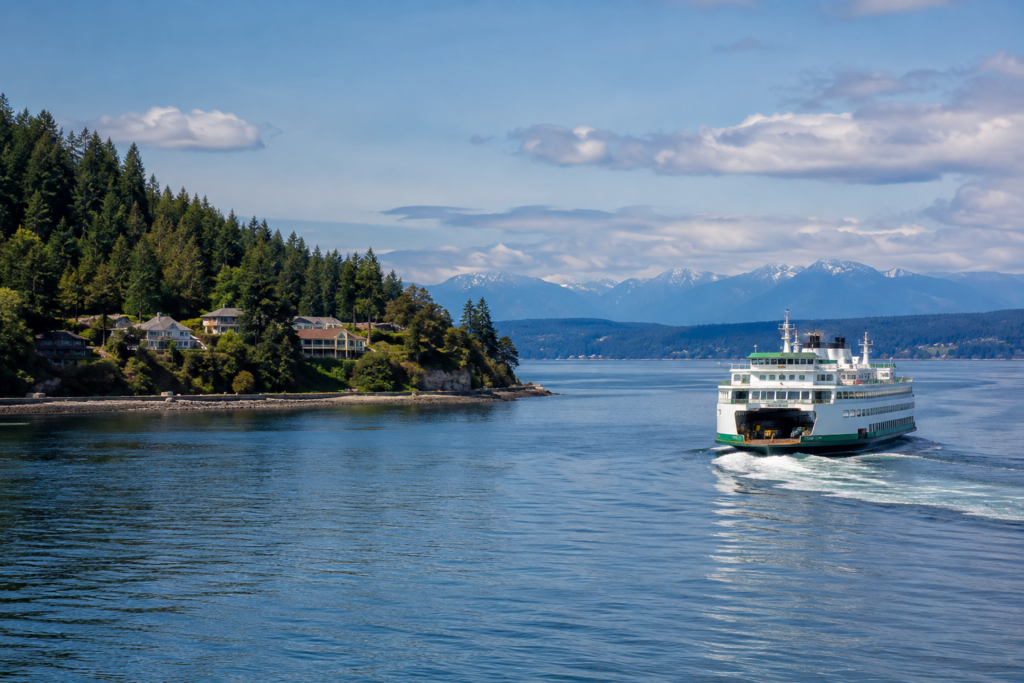 Águas calmas do Puget Sound com uma balsa navegando entre uma costa arborizada e montanhas ao fundo em um dia parcialmente nublado.