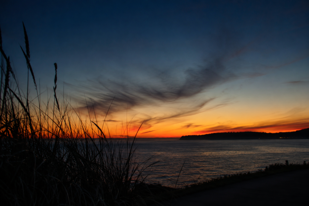 Vista ampla do litoral do Puget Sound ao entardecer, mostrando a longa claridade de verão refletida sobre a água sob um céu com tons de azul e laranja.
