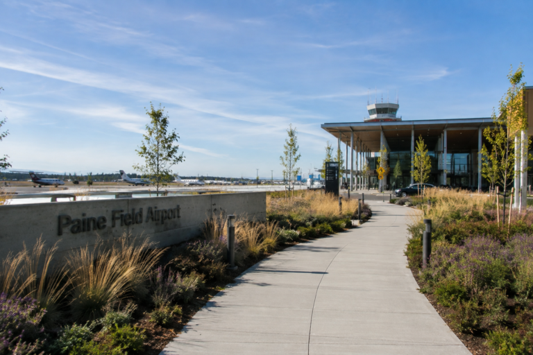 Caminho de pedestres em direção ao terminal de Paine Field, em Everett, com aeronaves e instalações do aeroporto ao fundo.