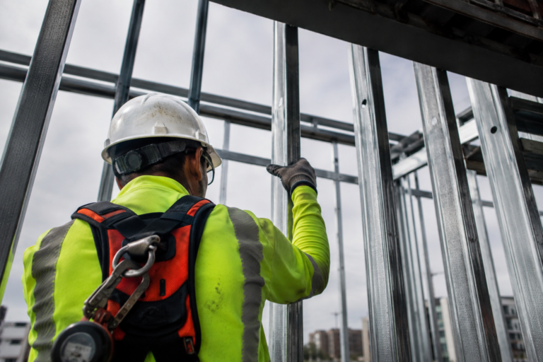 Trabalhador da construção civil visto de costas usando capacete, roupa de alta visibilidade e equipamento de segurança entre estruturas metálicas de uma obra.