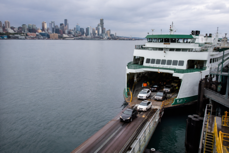 Balsa transportando carros em terminal à beira d’água, com o centro de Seattle ao fundo sob céu nublado.