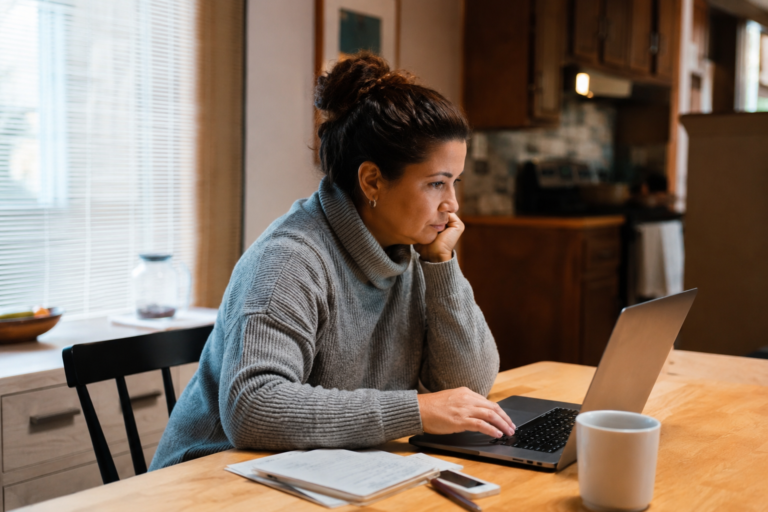 Mulher usando um laptop em uma mesa de casa, com papéis e celular ao lado, representando o acesso online a informações e opções de cobertura de saúde no estado de Washington.