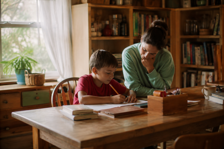Criança estudando em casa à mesa enquanto recebe orientação de uma adulta em um ambiente doméstico.