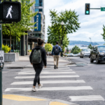Pedestres atravessando uma faixa de pedestres em um cruzamento urbano de Washington, com sinal de caminhada acionado e veículos circulando pela via.