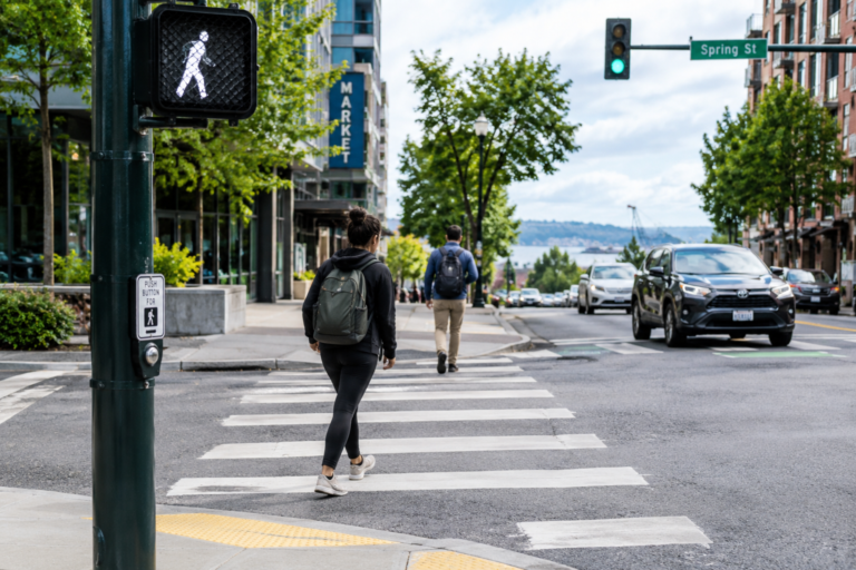 Pedestres atravessando uma faixa de pedestres em um cruzamento urbano de Washington, com sinal de caminhada acionado e veículos circulando pela via.