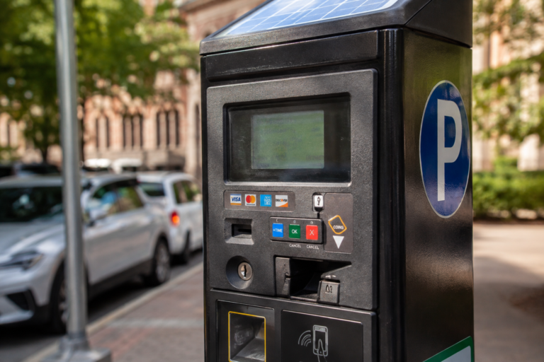 Parquímetro eletrônico em uma rua urbana, com carros estacionados ao fundo.
