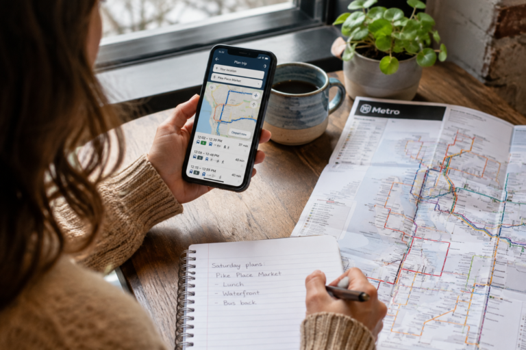 Pessoa planejando uma viagem de transporte público com celular, mapa de rotas e anotações sobre uma mesa.