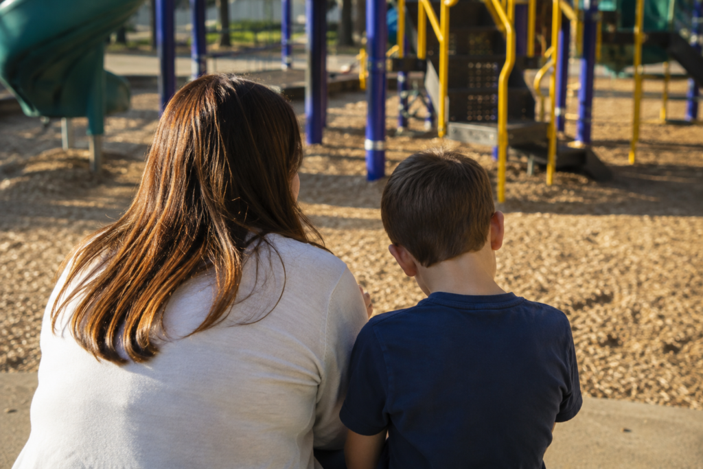 Mãe e criança vistas de costas em um parquinho escolar ao ar livre.