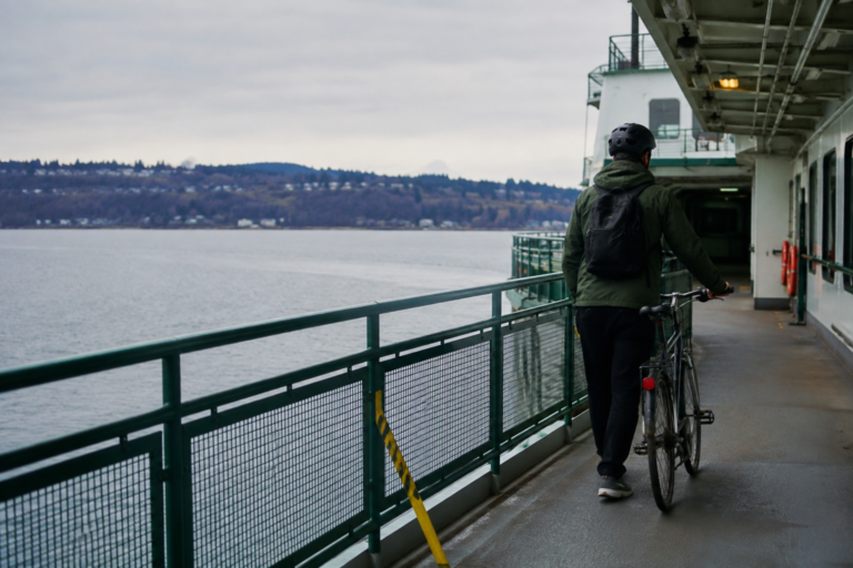 Ciclista conduzindo uma bicicleta pelo convés de uma balsa no Puget Sound, com água e costa arborizada ao fundo.