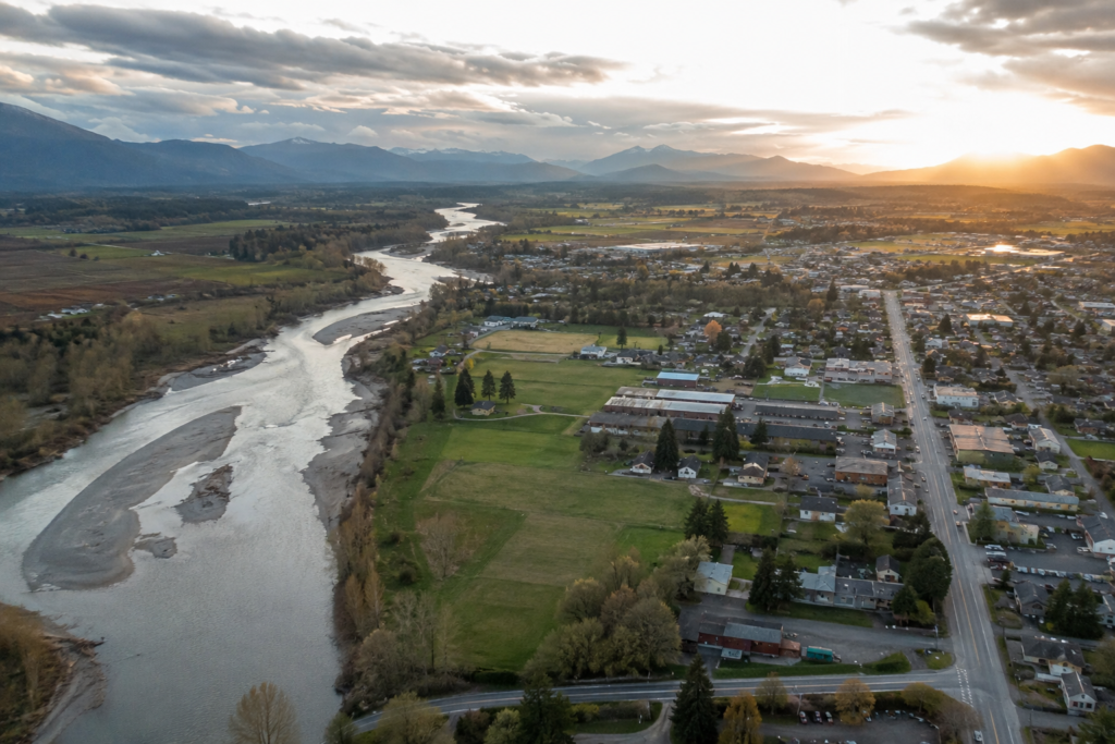 Vista aérea de Arlington, Washington, mostrando o rio Stillaguamish atravessando o vale, com bairros da cidade, áreas agrícolas e o sopé das Cascade Mountains sob a luz quente do fim do dia.