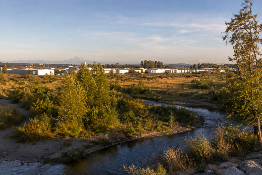 Vista elevada do Green River Valley em Kent, mostrando o rio em primeiro plano, áreas naturais, parques industriais ao fundo e o Monte Rainier no horizonte sob luz dourada do fim da tarde.