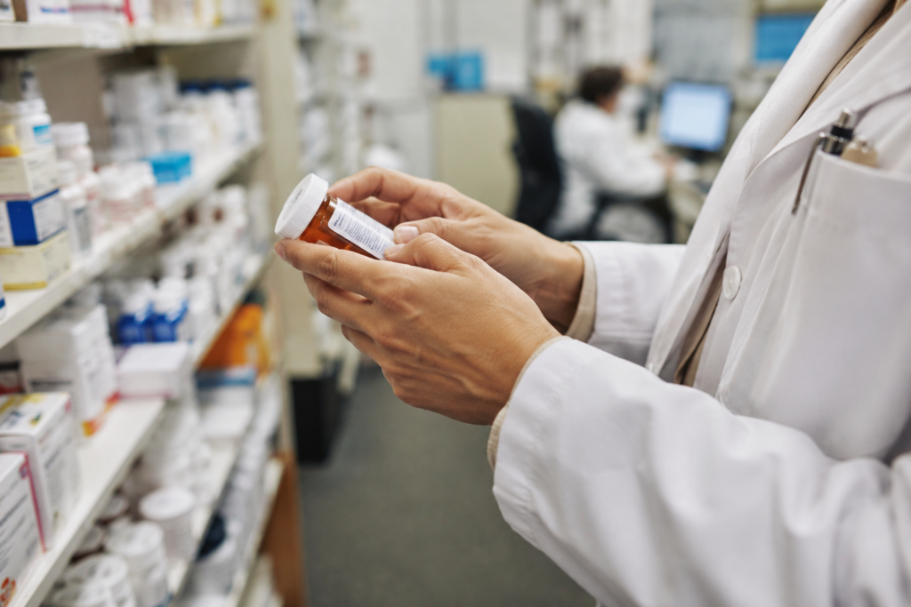 Farmacêutico segura um frasco de medicamento prescrito em uma farmácia, com prateleiras de remédios desfocadas ao fundo.