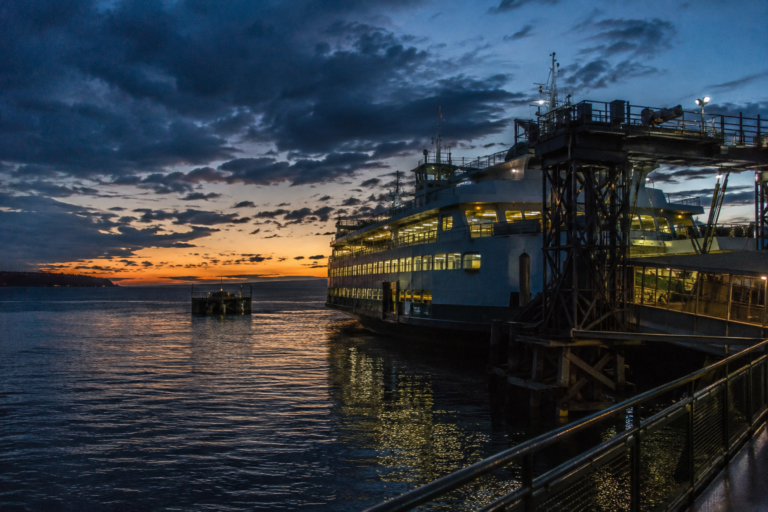Balsa atracada em terminal no Puget Sound ao entardecer, com infraestrutura portuária iluminada e águas da região ao fundo.