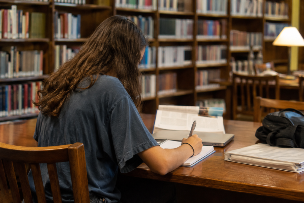 Estudante do High School estudando em uma biblioteca, com caderno, livros e materiais escolares sobre uma mesa.