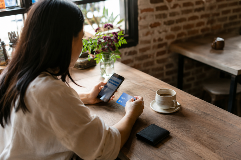 Mulher consultando informações bancárias no celular enquanto segura um cartão em uma mesa de café.