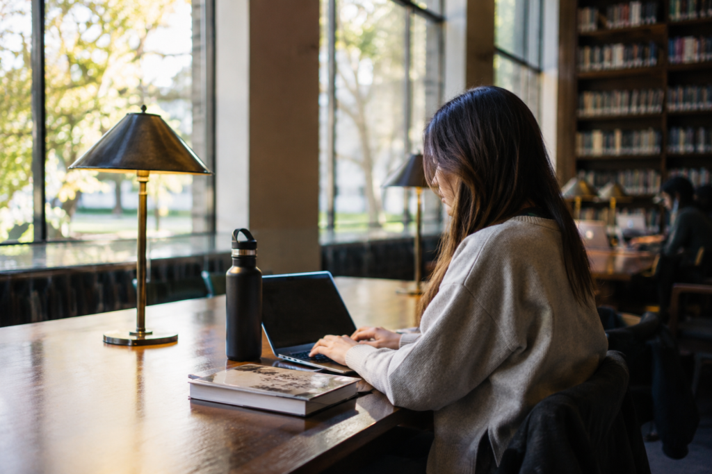 Estudante usando laptop em uma biblioteca universitária, com livros e materiais de estudo sobre uma mesa iluminada pela luz natural.