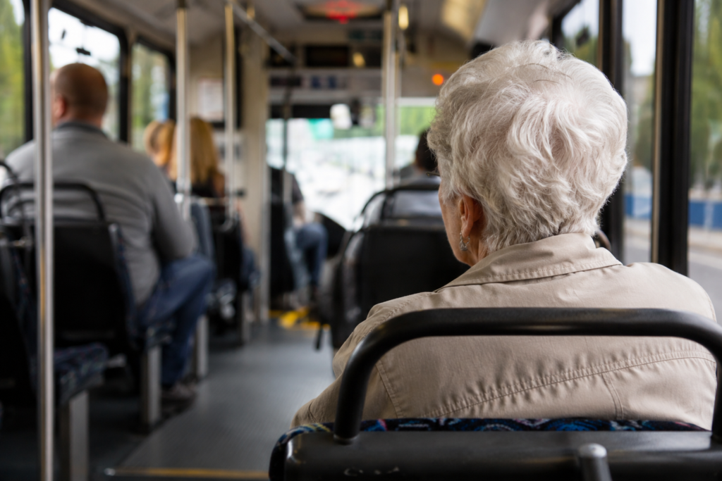 Passageira idosa sentada em um ônibus de transporte público, vista de costas, com outros passageiros ao fundo.